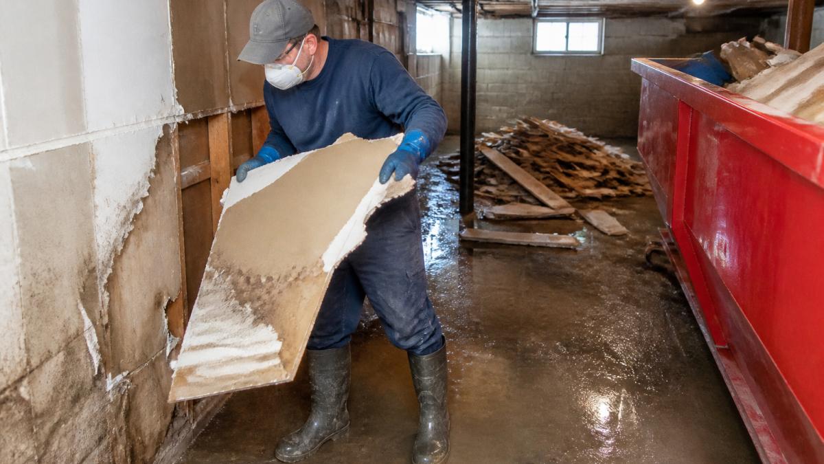 Homeowner wearing protective equipment including gloves, mask, and boots carefully removing water-damaged drywall from a flooded basement, with piled debris visible in background
